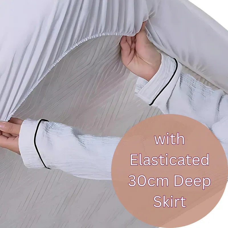 A close-up of a person pulling a white fitted sheet or pajama top toward themselves to demonstrate an elasticized, deep skirt. The person's hands and forearms are visible, and a peach-colored circular text overlay reads 'with Elasticated 30cm Deep Skirt'.